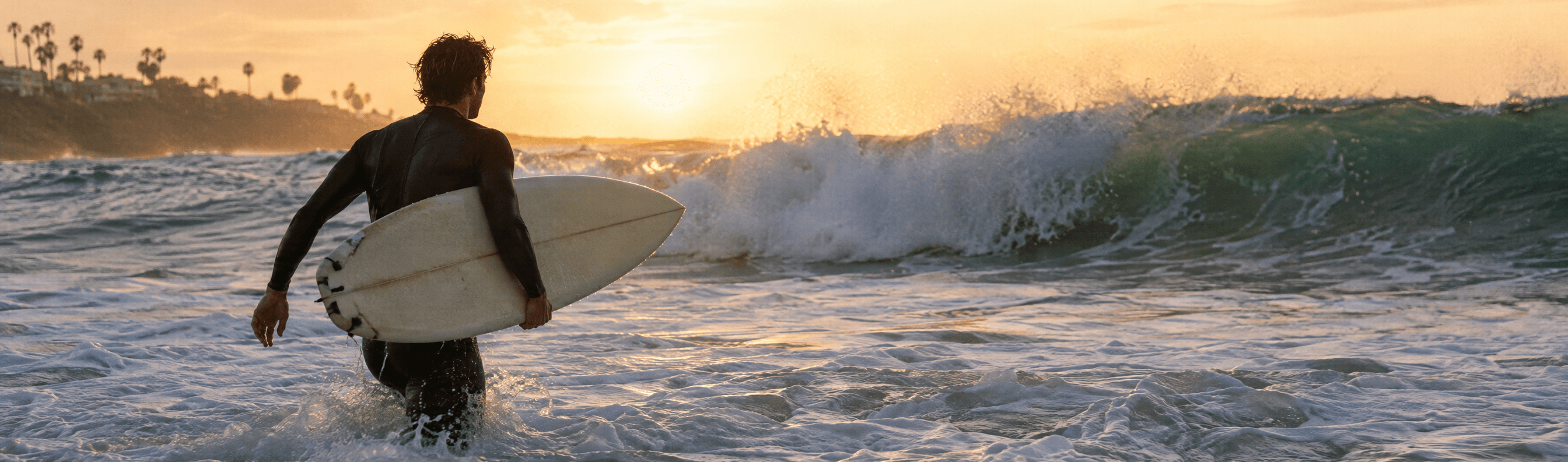 A surfer in a wetsuit wades into the whitewater at golden hour with a shortboard under his arm.